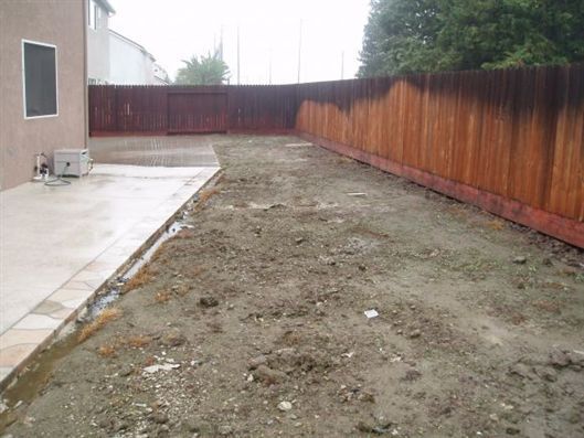 Narrow muddy backyard beside a house, with a concrete patio and a wooden fence on the right.