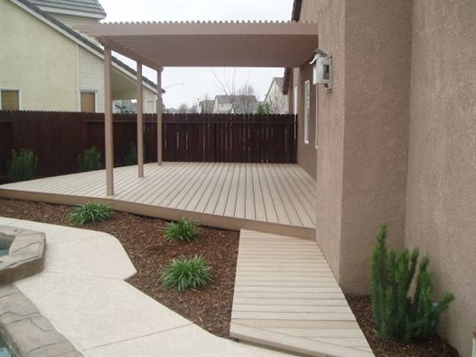 Covered backyard deck with wooden steps, patio pavers, and a brown fence beside a house.