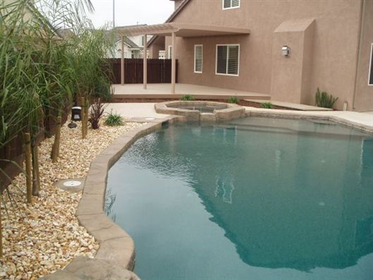 Backyard swimming pool with stone edging beside a stucco house and gravel landscaping