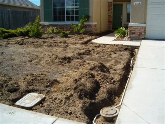 Bare dirt front yard beside a concrete walkway and house entrance, with landscaping drain covers.