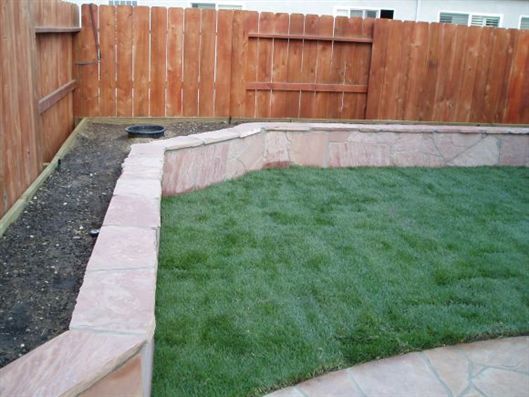 Backyard with a curved stone retaining wall, green grass, and a wooden fence.