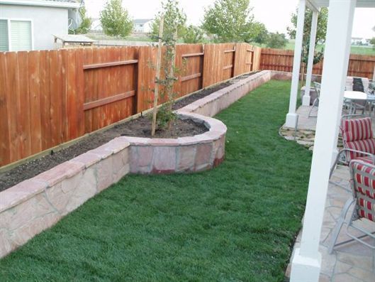 Backyard with curved raised planter wall, grass lawn, wooden fence, and patio chairs