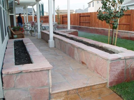 Stone patio with raised pink planters beside a covered walkway and fenced backyard grass.