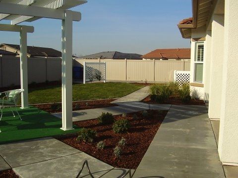 Backyard patio and walkway beside a house, with pergola, lawn, and fenced yard under clear skies