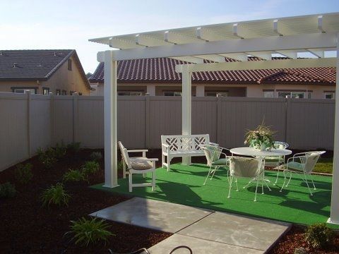 Backyard patio with white pergola, green turf, and outdoor table and chairs under a cloudy sky