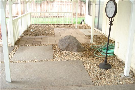 Covered porch with gravel and paving stones, a hose reel, and a black standing lamp.