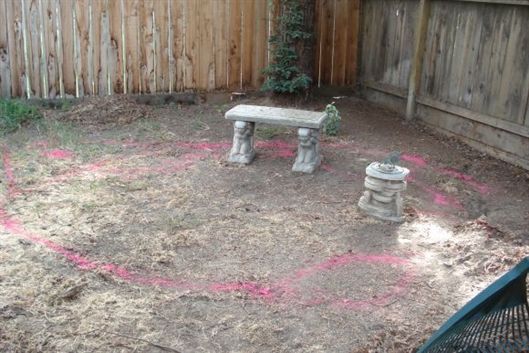 Backyard dirt area outlined in pink with a small stone bench and pedestal near a wooden fence.