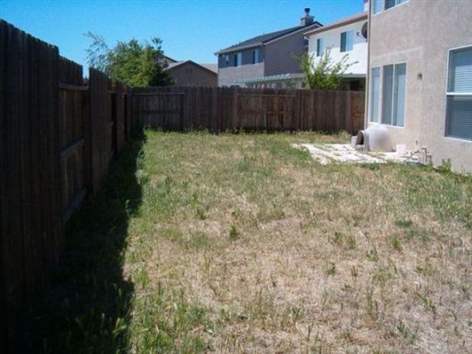 Narrow backyard with patchy grass, wooden fence on left and back, and a house wall on the right.
