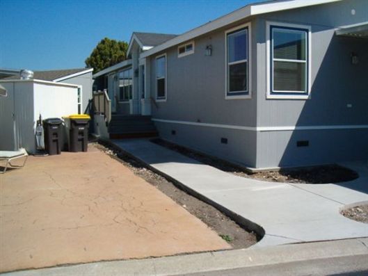Gray manufactured home with a concrete ramp and sandy yard beside trash bins under a clear blue sky