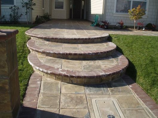 Curved stone steps leading up to a front patio and house entrance, bordered by grass and brick edging.