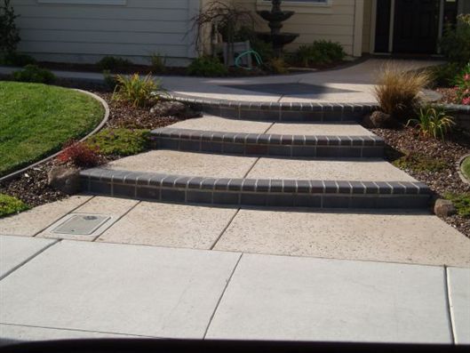 Front steps with three concrete stair tiers leading to a house entrance, bordered by landscaping and sidewalk