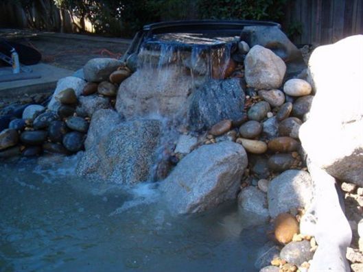 Rock waterfall cascading over stacked stones into a blue pond in a landscaped garden