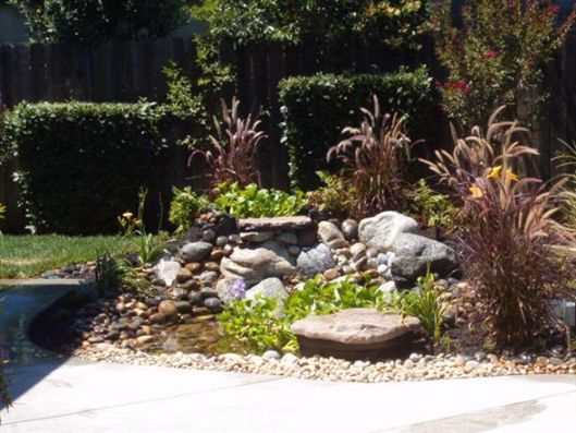 Landscape rock garden with grasses, white stones, and a stepping stone beside a patio and hedge backdrop