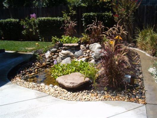 Rock garden with stones, plants, and a small pond beside a concrete patio.