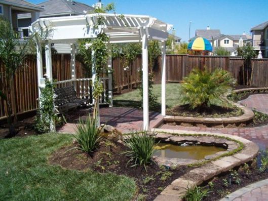 Backyard garden with white pergola, wooden fence, pond, and landscaped plants