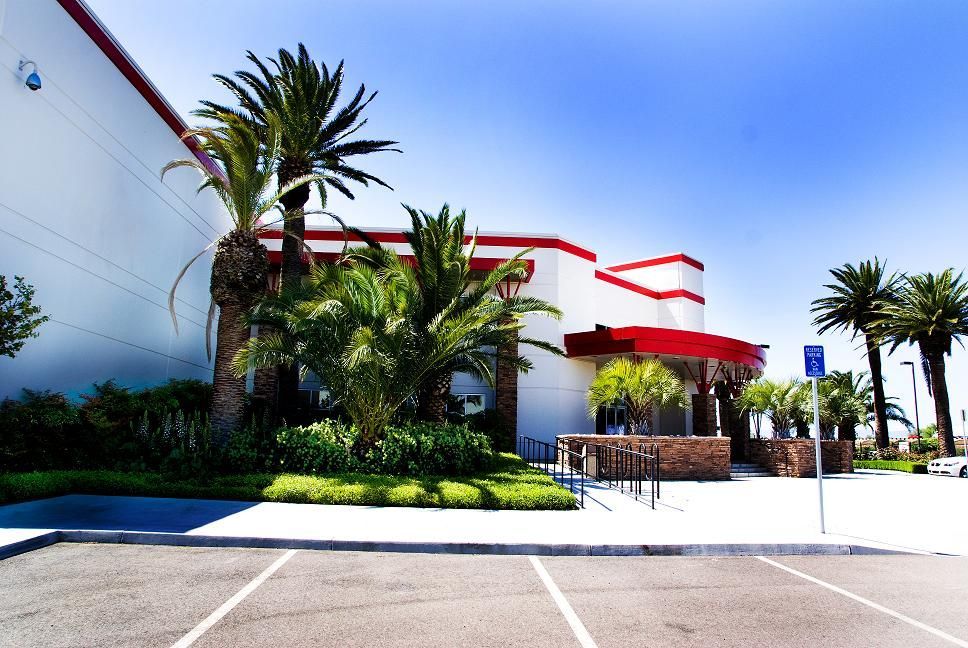 Modern building entrance with red accents, palm trees, and landscaped greenery under a bright blue sky