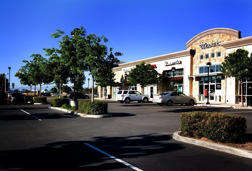 Sunny shopping center strip mall with parked cars, trees, and storefronts under a blue sky