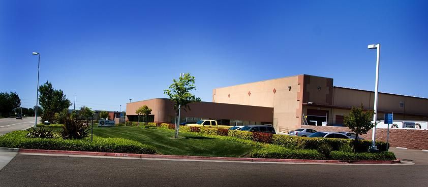 Large tan commercial building with red shrubs and parking lot under a clear blue sky