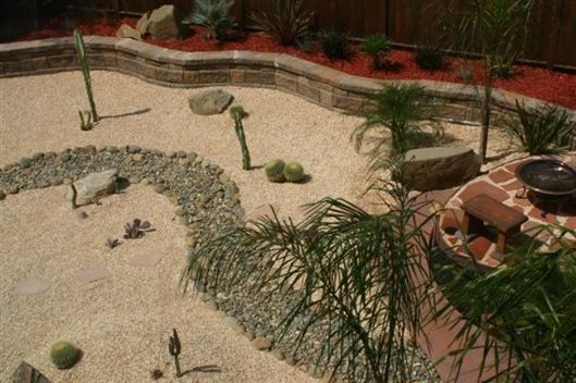Desert-style backyard with gravel, rocks, cacti, and a stone fire pit with chairs.