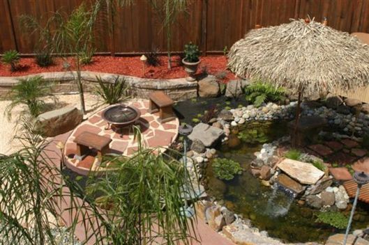 Backyard pond with rocks, plants, and a thatched umbrella beside a brick patio