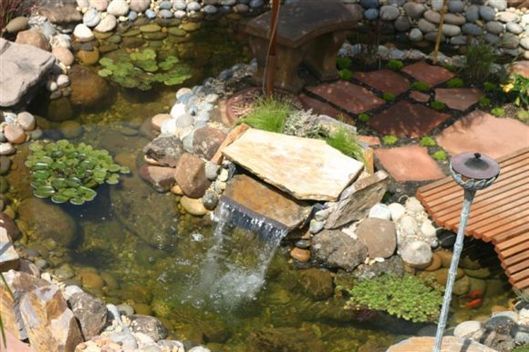 Small rock pond with a trickling waterfall, lily pads, and a garden lamp beside a stone border.