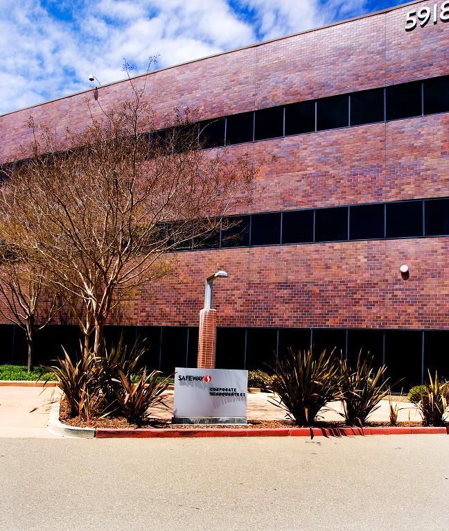 Brick office building with dark windows, leafless tree, desert landscaping, and a sign by the curb.