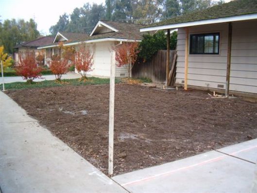 Bare yard plot beside a house, with a concrete walkway and a white post in the foreground.