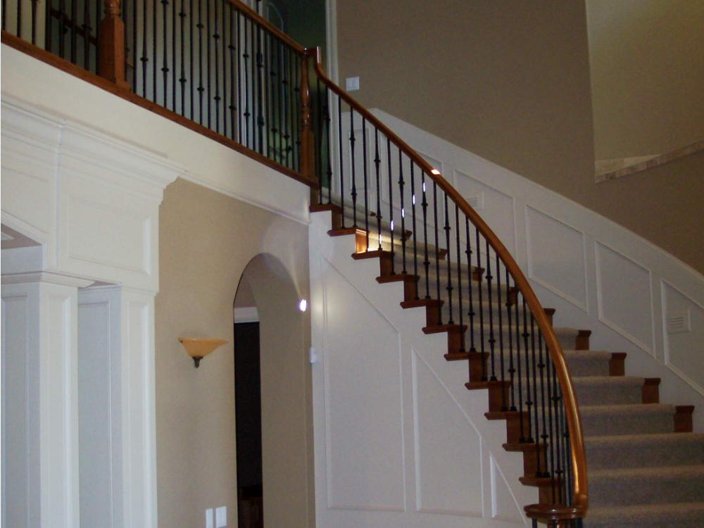 A curved staircase with wooden steps and railings, black metal balusters, and white wainscoting on the wall.