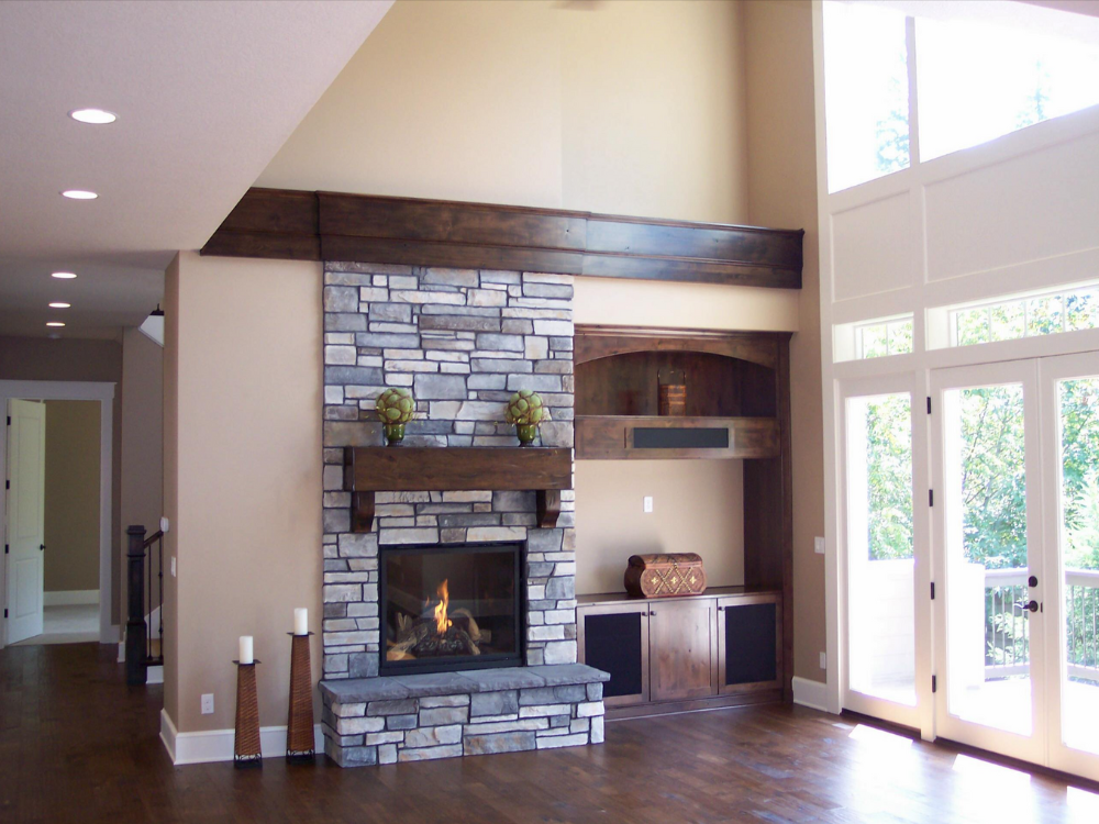A living room with a floor-to-ceiling stone fireplace, dark wood mantel, built-in shelving, and large windows.