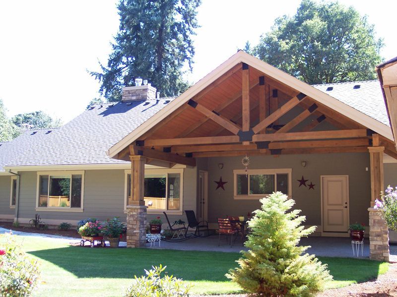 A covered patio addition with a wooden truss roof, stone pillars, and lawn, attached to a gray house with a shingled roof.