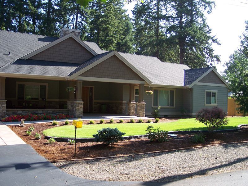 A one-story tan house with a covered porch, stone accents, and a manicured green lawn in front of a forest.