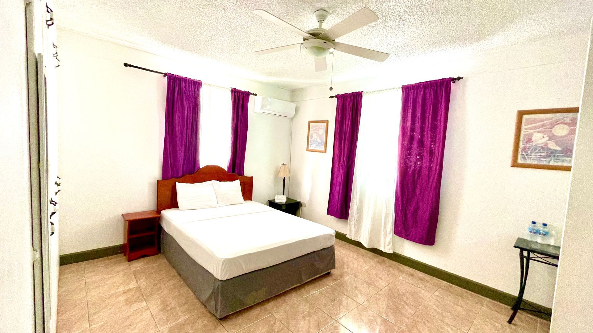 A bedroom with a bed , nightstand , ceiling fan and purple curtains.
