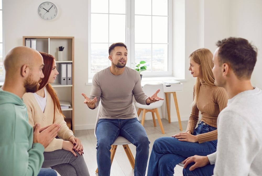 Five people sit in a circle in a bright office, conversing as part of a group meeting or therapy session.