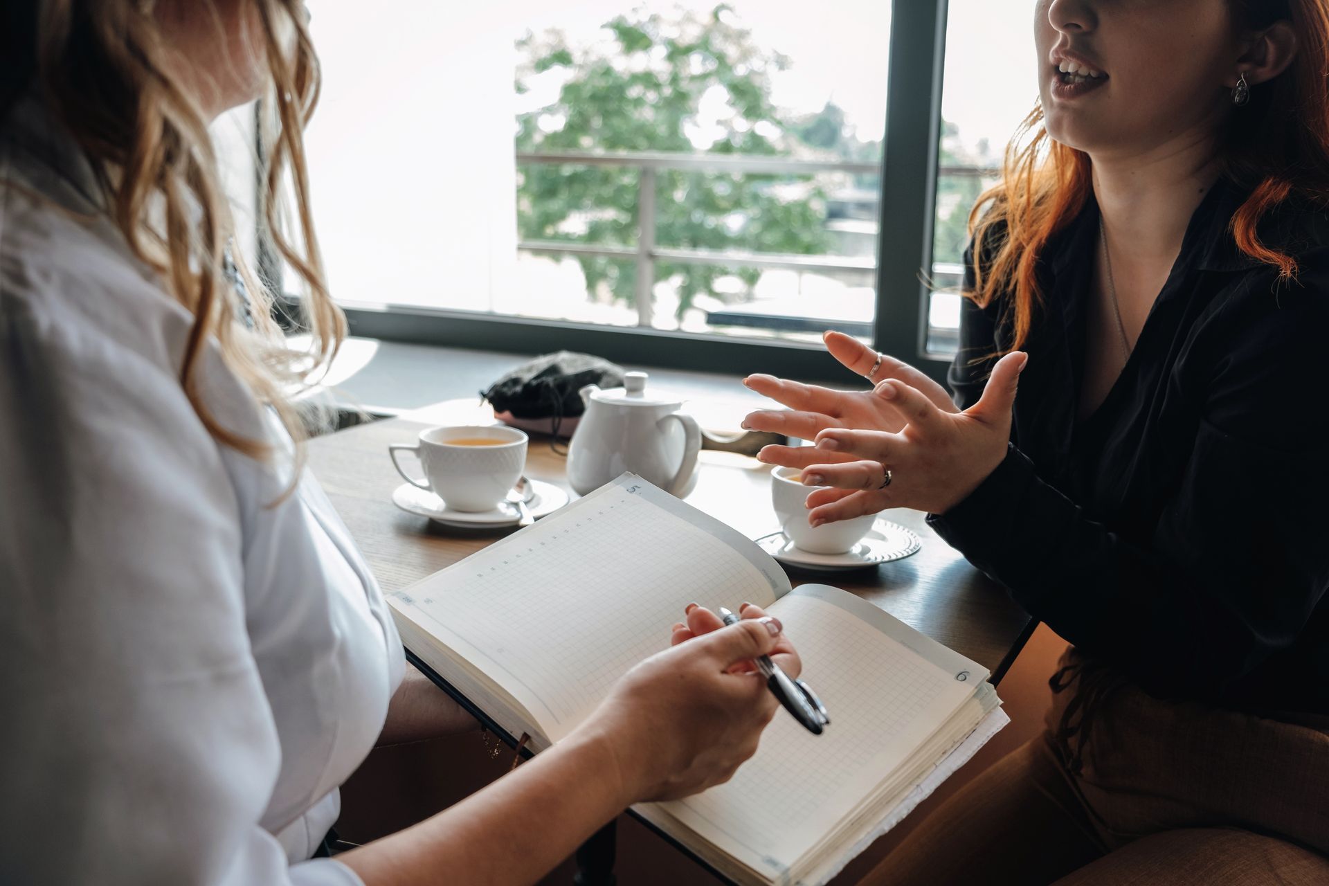 Two people sit at a table with drinks and an open notebook, one speaking while the other prepares to write.