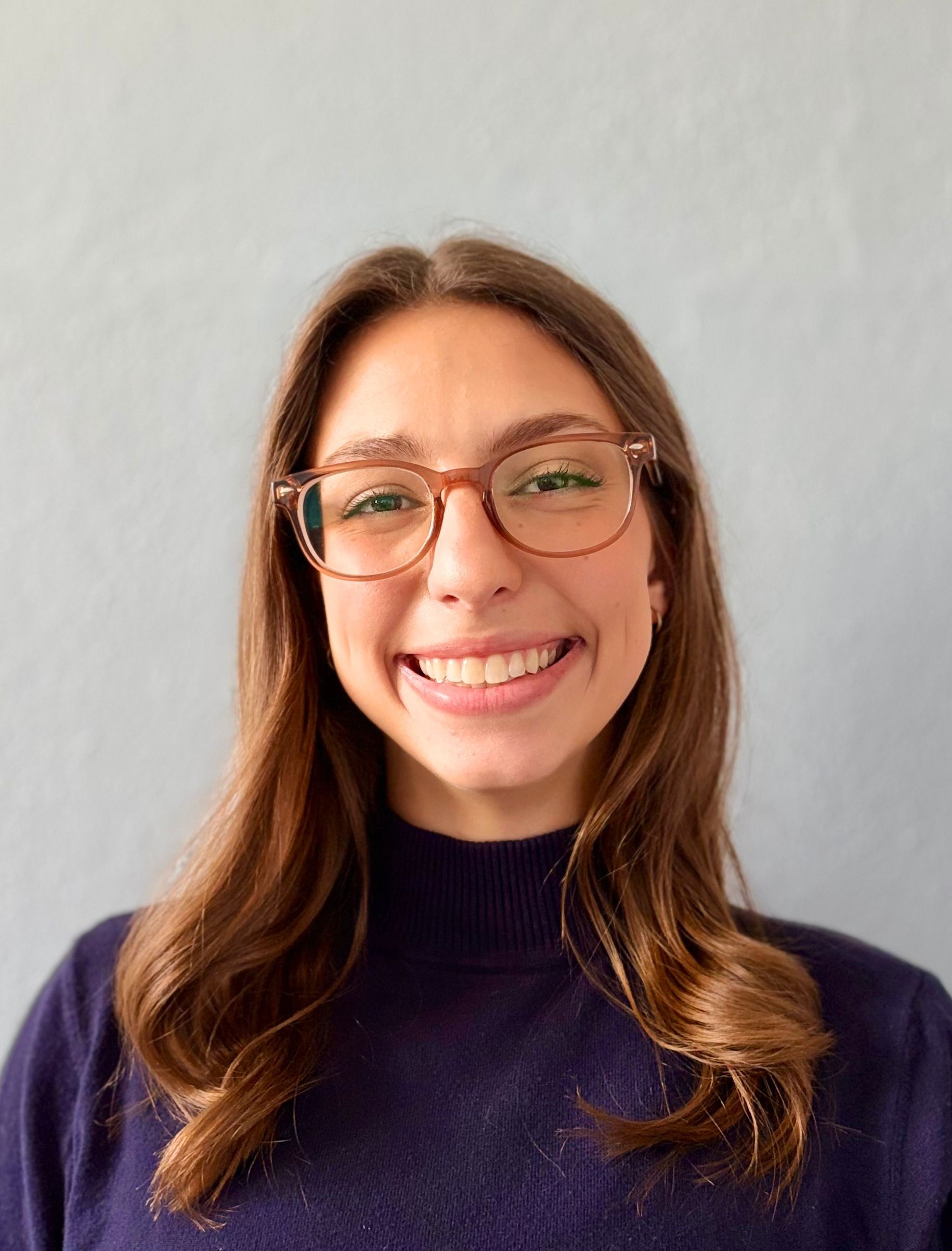 A person with long brown hair and dark-rimmed glasses smiling at the camera in an indoor setting.