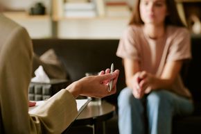 A professional sits with a clipboard and pen, facing a person sitting on a sofa in a therapy or counseling session.