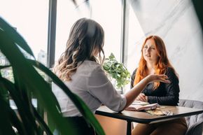 Two people sit at a table by a window in a sunlit room; one is speaking with a hand gesture while the other listens.