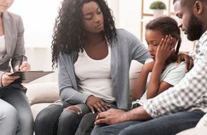 A family sits with a professional, with parents comforting their young child who is holding their head.