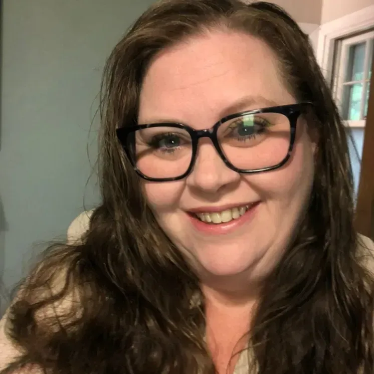 A person with long brown hair and dark-rimmed glasses smiling at the camera in an indoor setting.
