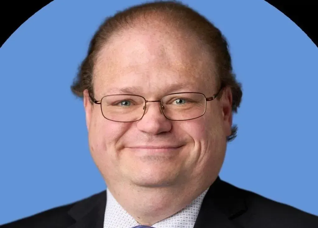 Headshot of a man with light brown hair wearing glasses and a dark suit against a solid blue background.