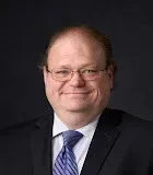 Headshot of a smiling person wearing glasses, a white collared shirt, blue striped tie, and a dark suit against black.