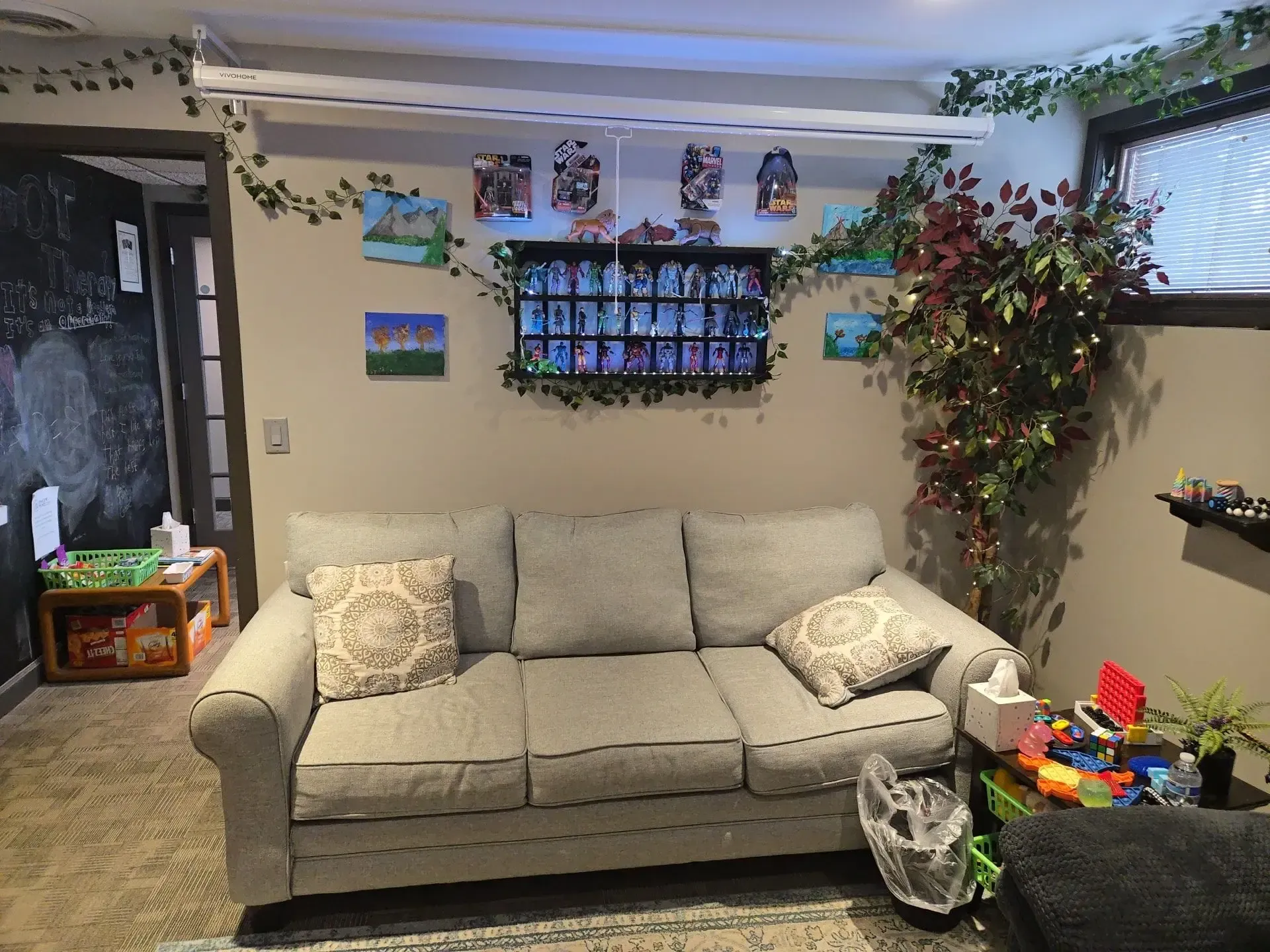 A beige couch sits in a room decorated with fake vines, small paintings, a display shelf, and an artificial tree.