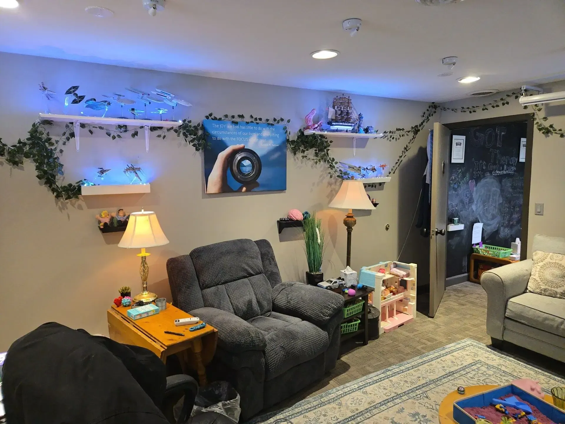 A cozy living room with a recliner, lamps, wall shelves with vines and blue lights, and an open doorway to a dark room.