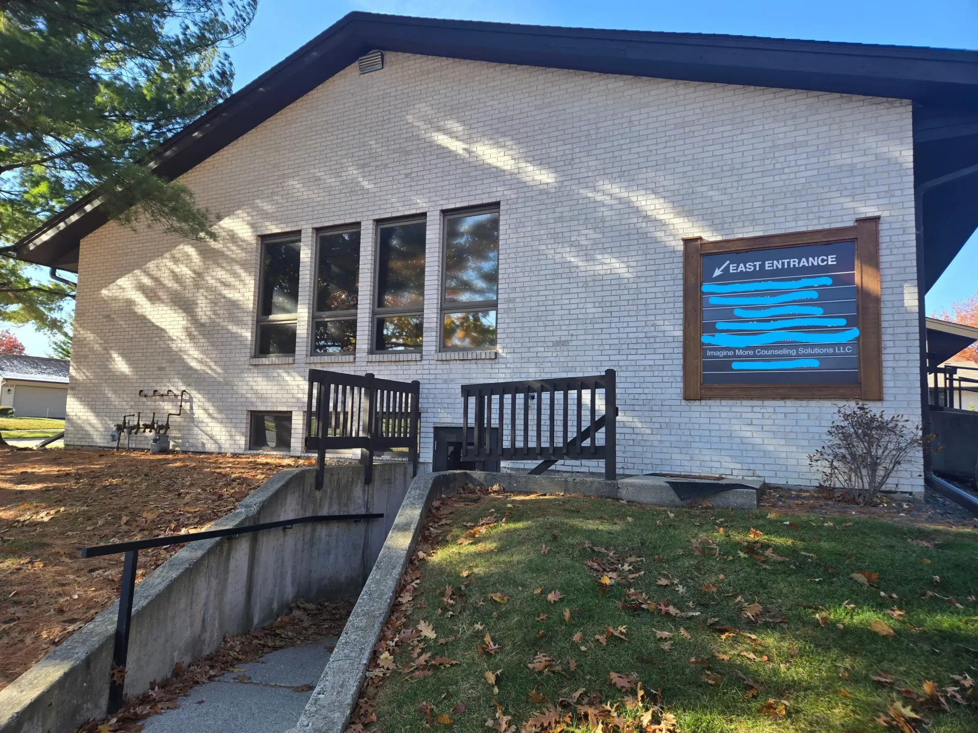 A white brick building with a blue sign, a small railed deck, and a concrete ramp leading to a basement entrance.