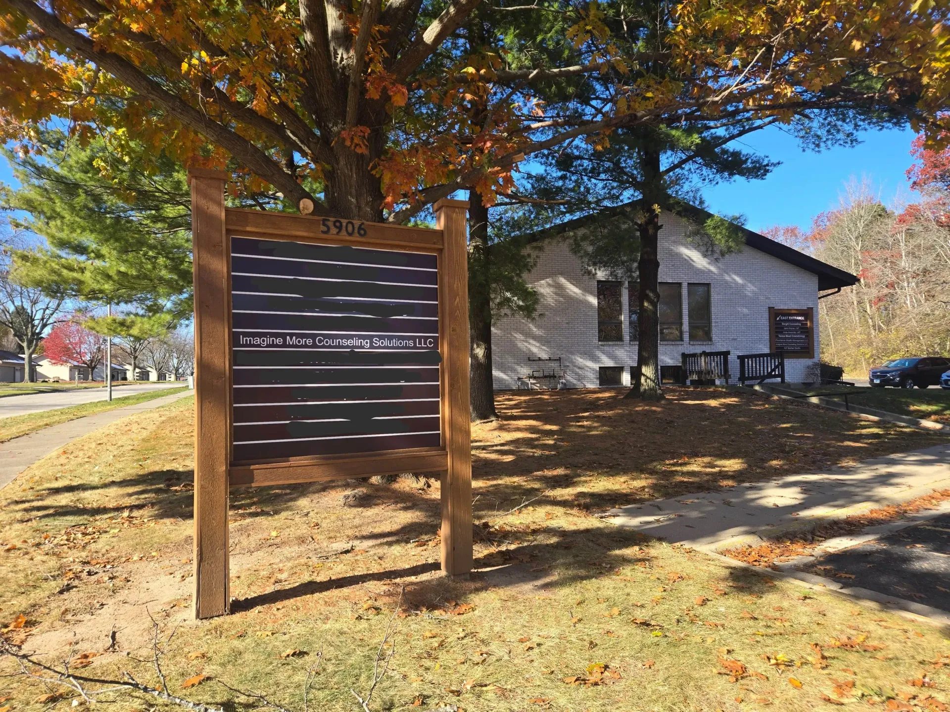 A brown wooden sign board stands on a lawn in front of a white building on a sunny autumn day with fallen leaves.