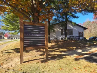 A brown wooden sign with missing letters stands in front of a white house with trees in an autumn landscape.