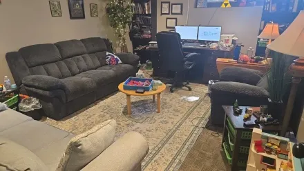 A cozy living room with a dark grey sofa, a light grey couch, a coffee table, and a dual-monitor computer desk setup.