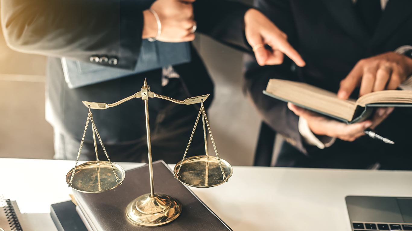 Two lawyers, one pointing at a law book, next to a balance scale, on a desk.