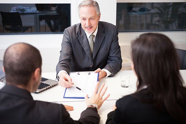 A man in a suit and tie is sitting at a table talking to two people.