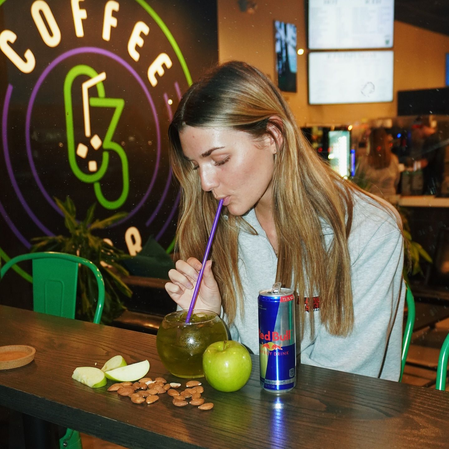 Woman drinking from a glass, sitting at a bar with an apple and a can of Red Bull.